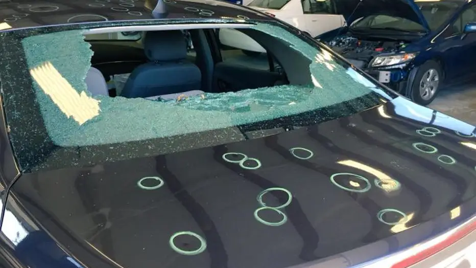 Sedan in a repair shop with a shattered rear windshield and glass fragments on the hood among other cars.