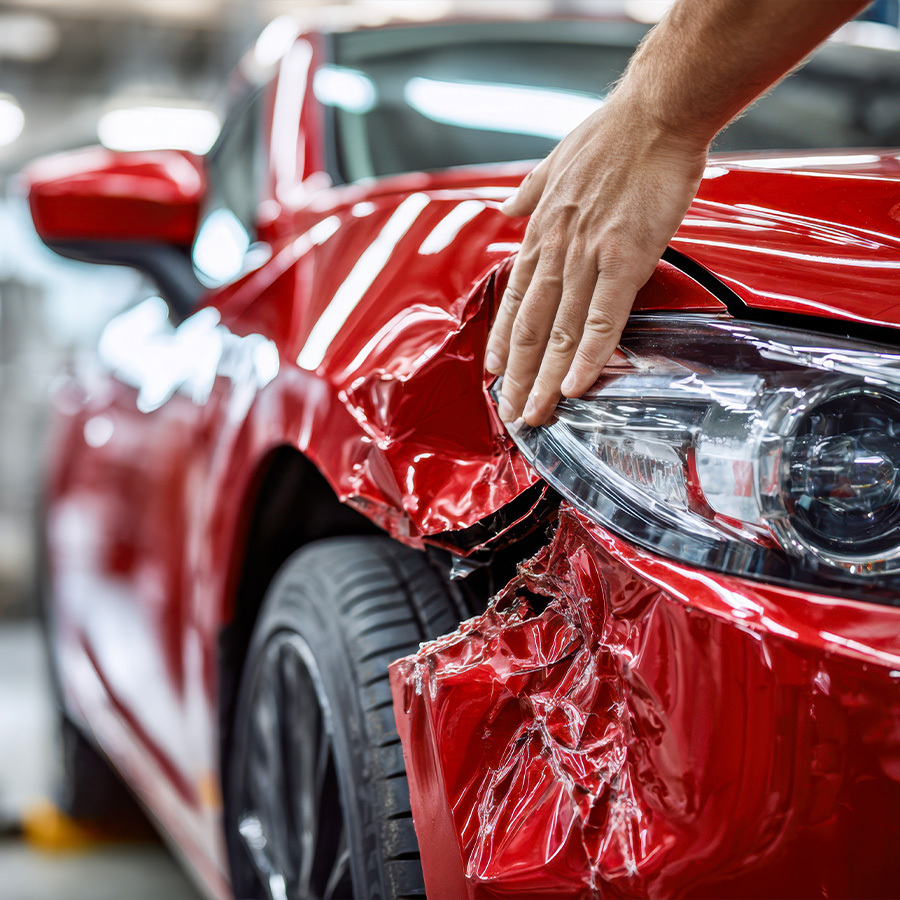 Close-up of a mechanic's hand on a damaged red car front fender in a repair shop.