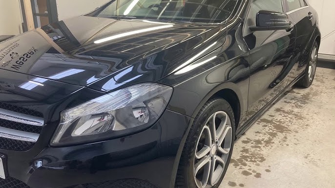 Front-left view of a black car parked indoors, showing the headlight, grille, and alloy wheel with reflections on the hood surface.