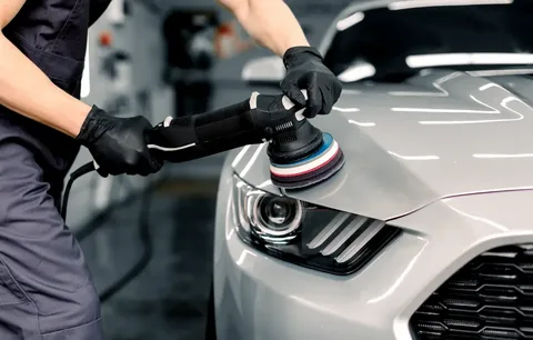 Auto technician wearing gloves polishes the front of a silver car with a rotary polisher.