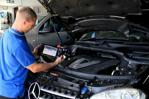 Auto mechanic in a blue uniform using a handheld diagnostic tablet under the open hood of a car in a workshop.