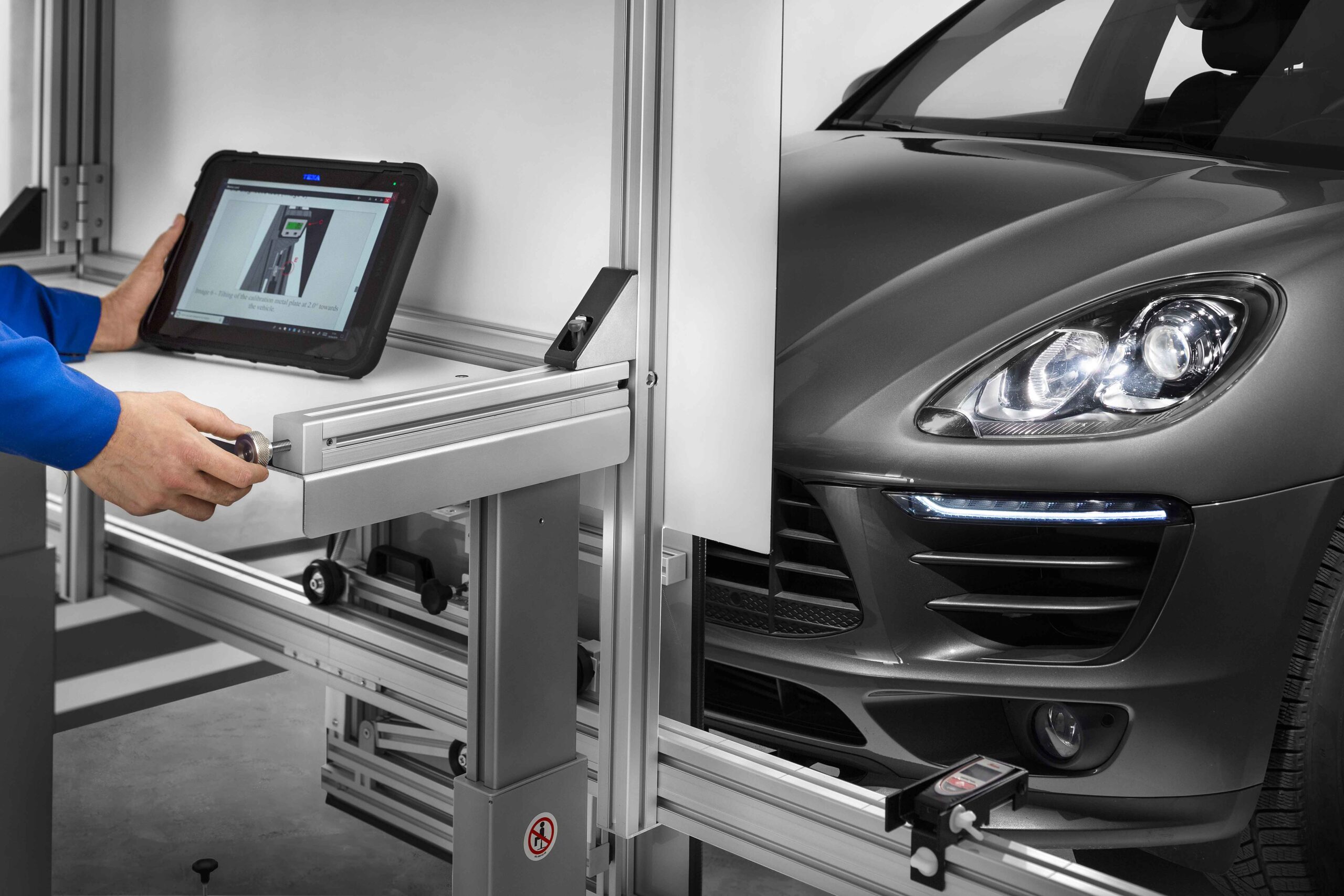 Technician using a diagnostic tablet on a vehicle testing rig beside a gray car inside a workshop stall.