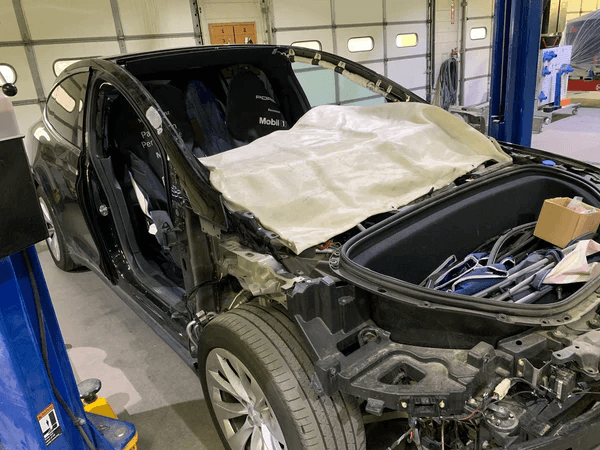Black coupe in a repair shop with the front end torn apart, hood area exposed and a white protective sheet draped over the cockpit.
