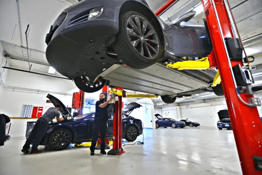 Auto repair shop with a gray car elevated on a hydraulic lift, a mechanic underneath, and another technician working on a blue car with its hood open in the background.