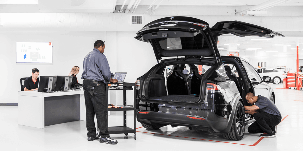 Tech kneeling by the rear wheel of a black Tesla with the hatch open in a bright service bay, while a mechanic works underneath the car.