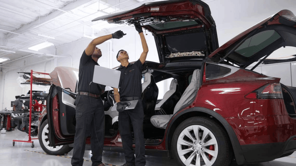 Two technicians inspect a red car with its trunk open, one holding a laptop and the other pointing at the engine area in a bright workshop falsely with white interior lights.