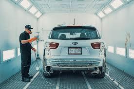 A mechanic inspects the rear of a white SUV inside a bright auto paint booth.