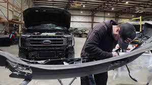 Mechanic wearing a cap, hood open, working on a vehicle with the front bumper removed in a garage set-up.
