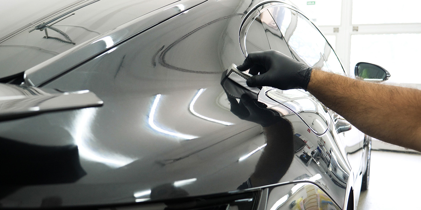 Technician wearing black gloves polishing a car's exterior with a cloth, reflecting bright workshop lights on a glossy surface.