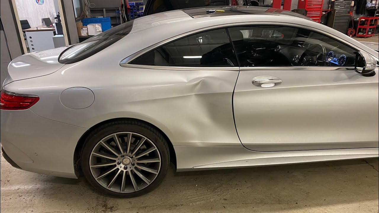 Silver luxury car in a workshop with a large dent on the rear door and mid‑section, showing a multi‑spoke wheel and service bay in background