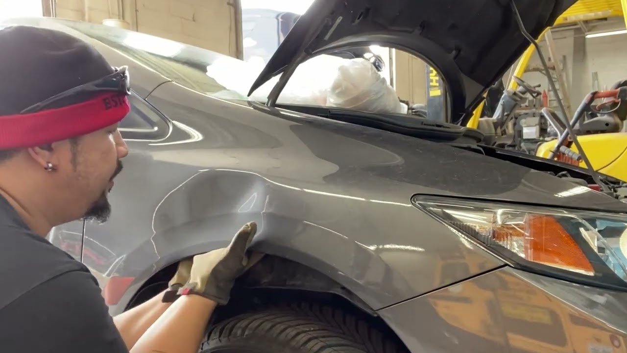 Male auto technician with a red headband and gloves repairs a dent on the front fender of a gray car with the hood open in a workshop.