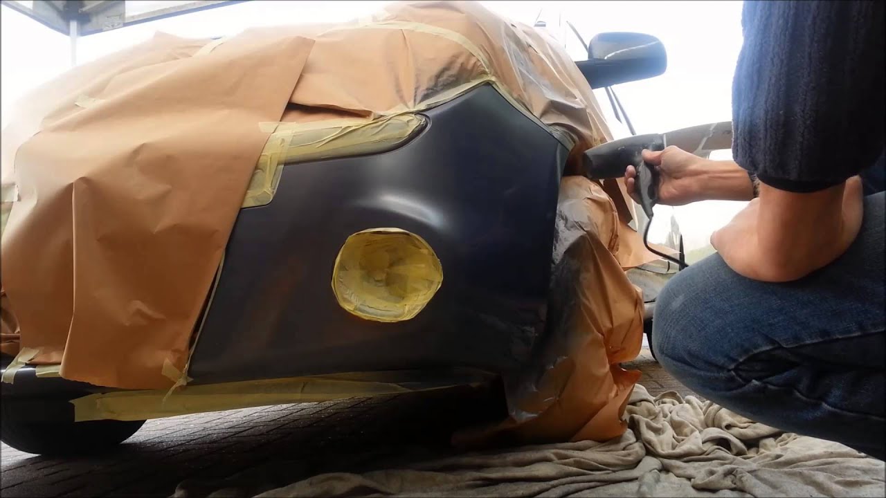Person spray-painting a car panel, masking paper and tape covering surrounding areas