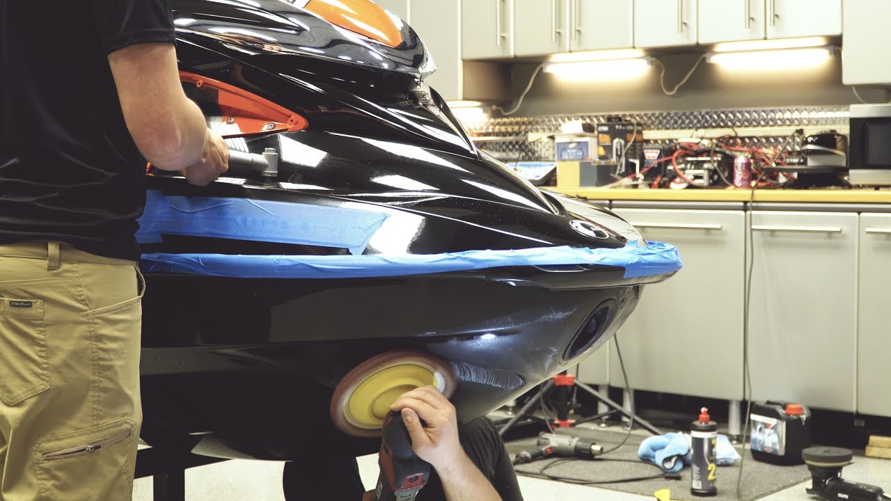 Technician polishing the rear of a glossy black car in a workshop, blue masking tape along the bumper.