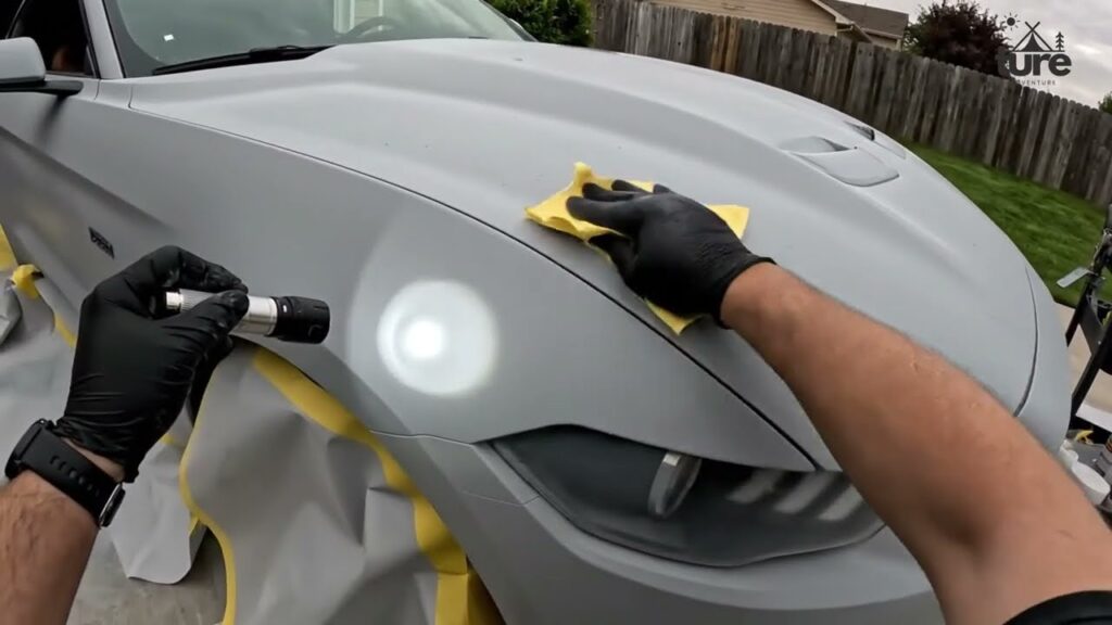 A person wearing black gloves polishes the front left fender of a gray car with a handheld buffer, masking tape visible around the area.