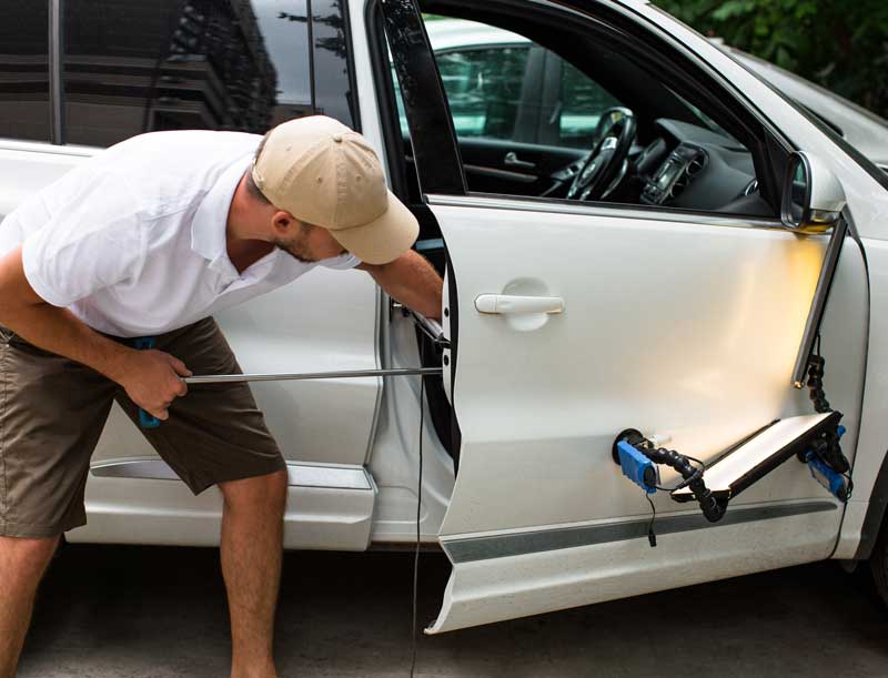 Man in a cap repairs a silver car door, tool in hand, with the door open
