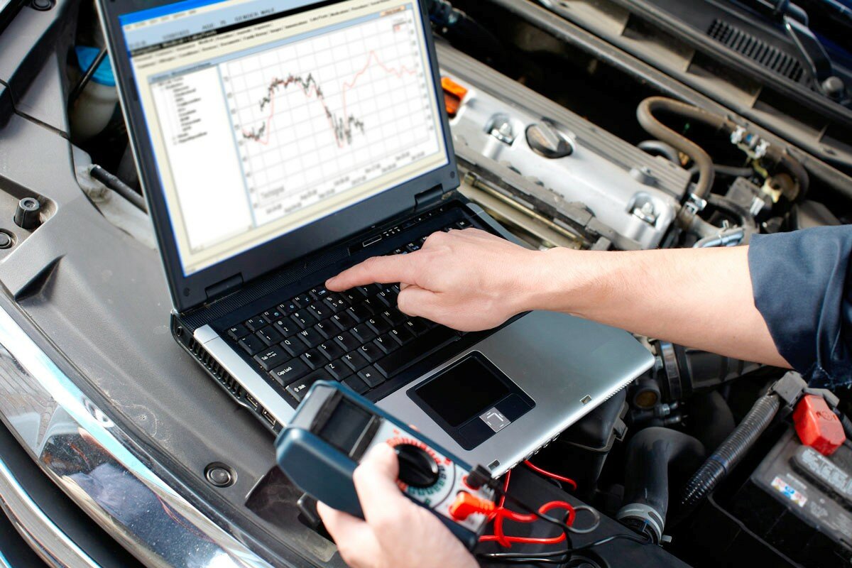 Mechanic using a laptop in the car engine bay to diagnose the electrical system, with a handheld multimeter connected nearby.