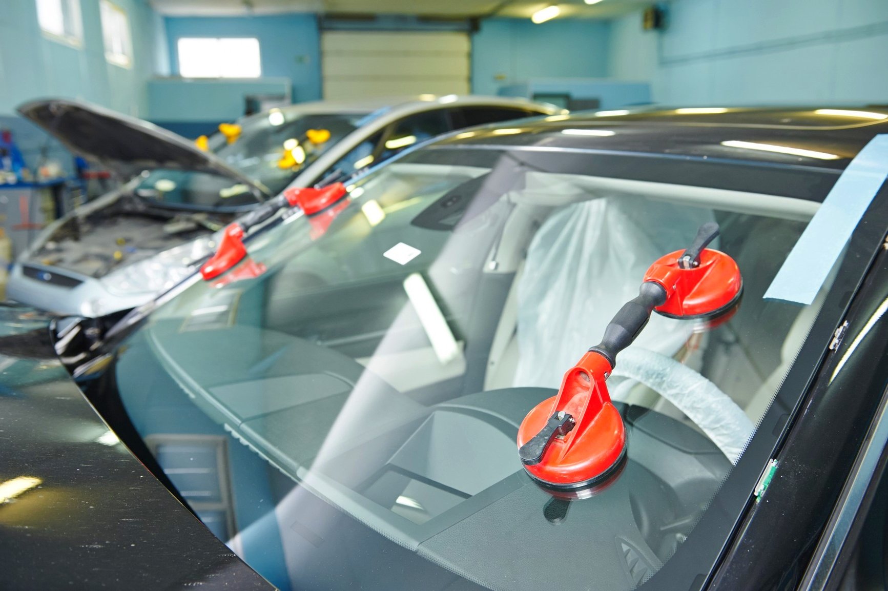 Inside an automotive repair shop with multiple cars, hoods open, and red suction cup tools attached to windshields indicating windshield work.
