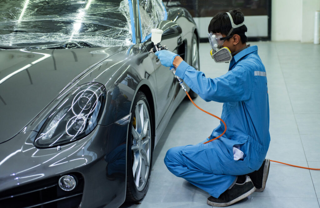 Auto body technician wearing a respirator and blue coveralls spray-paints a dark car door with a spray gun.