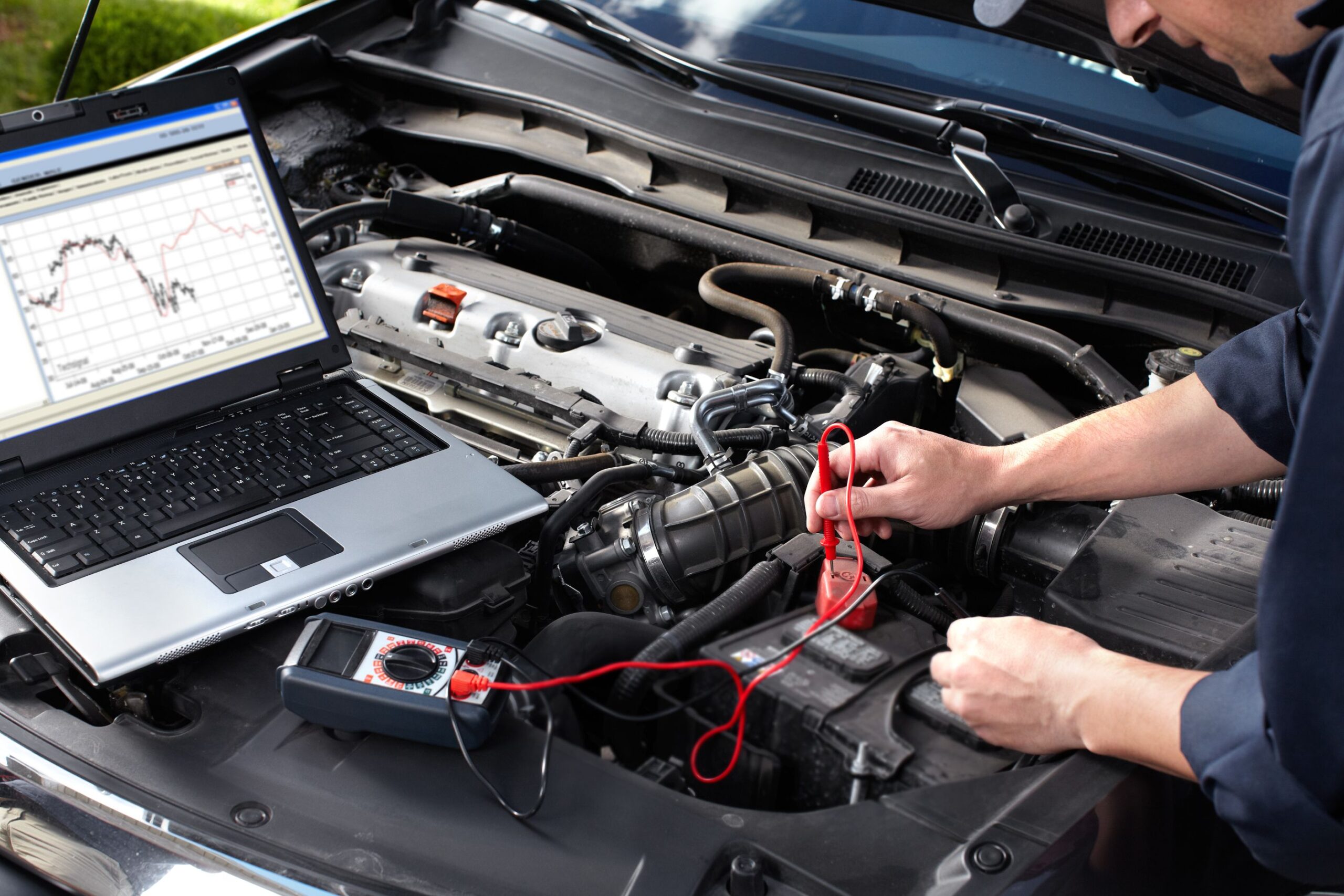 Technician using a laptop to diagnose a car engine with test leads connected under the hood of a vehicle.