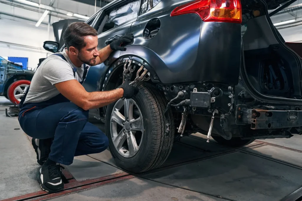 Auto technician kneels by a dark SUV, working on the front wheel with tools in a busy repair shop