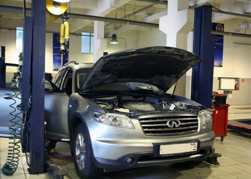 Silver Infiniti SUV with its hood up in a car repair shop, engine bay exposed and workshop equipment nearby.