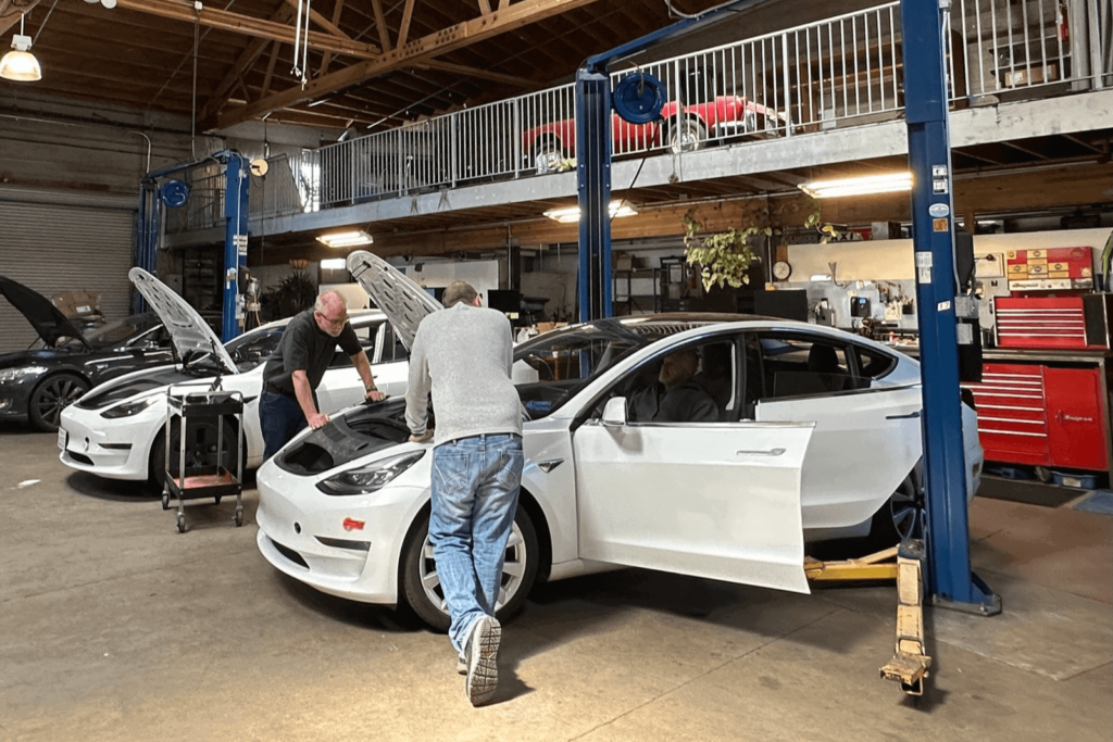 Two mechanics inspect the open hood of a white electric car inside an auto repair shop, with another white car nearby and a blue car lift overhead.