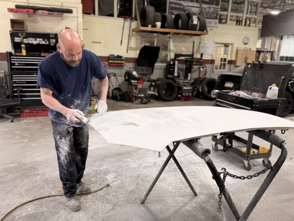 Bald man in a blue shirt sands a white car hood on a workbench in an auto body shop.
