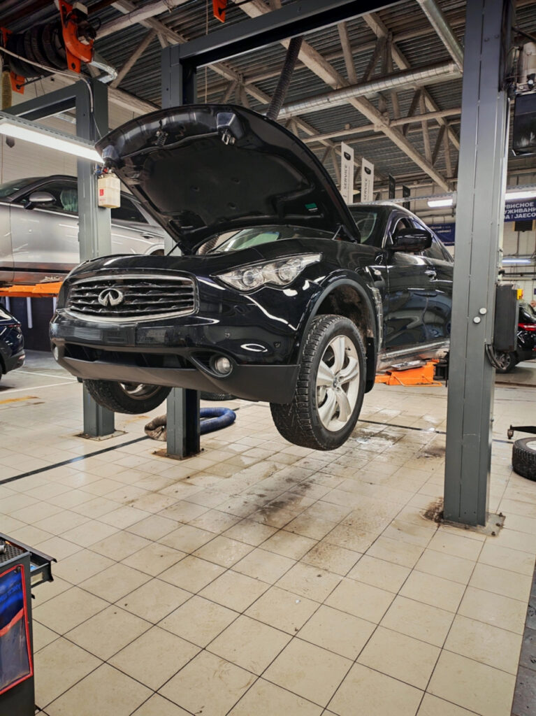 Black Infiniti SUV on a lift with its hood open in an auto repair shop, front wheels raised for service inspection.