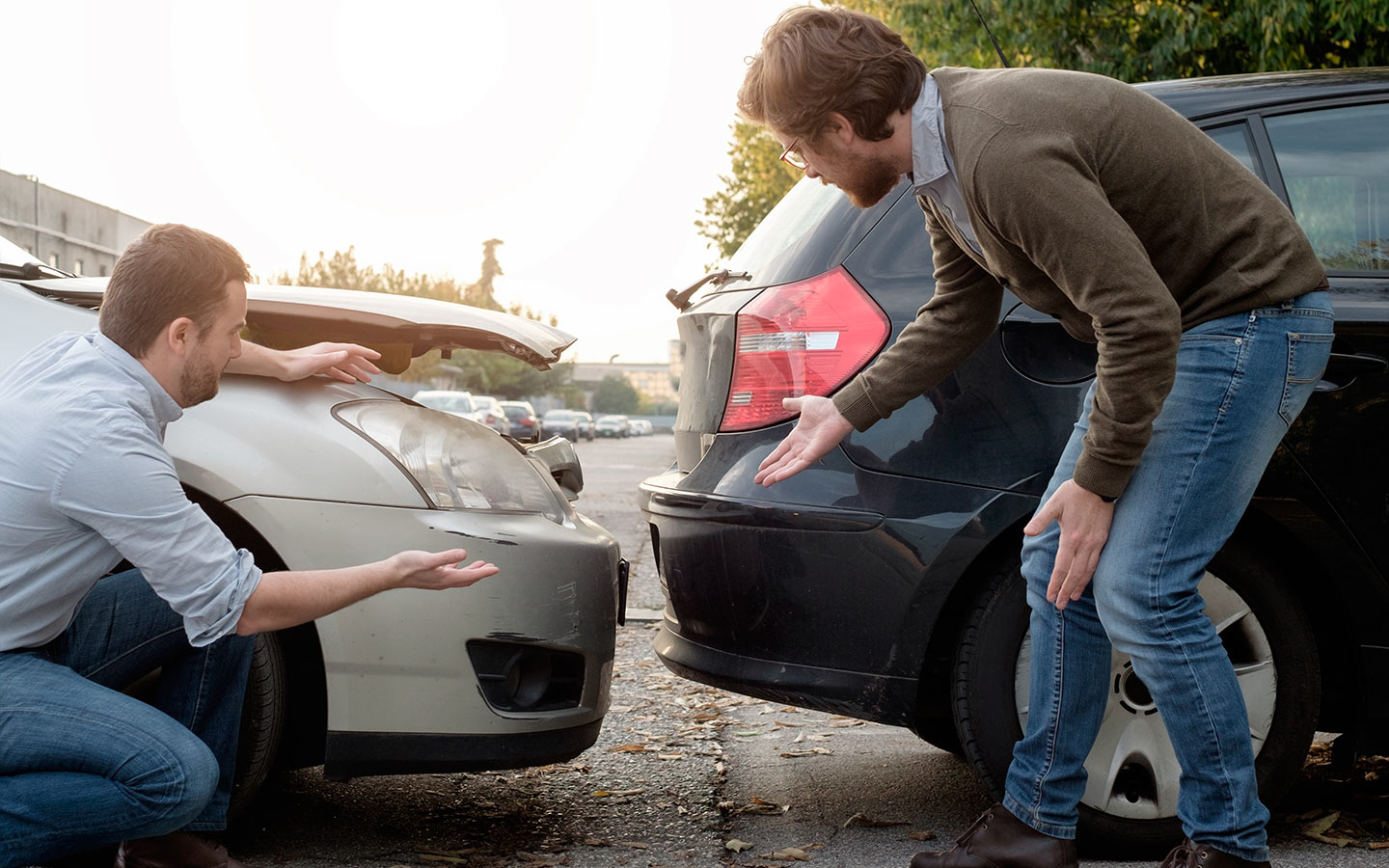 Two men assess damage after a car collision on a street, one crouching by a gray car with its hood up and the other pointing to the back of a dark SUV.